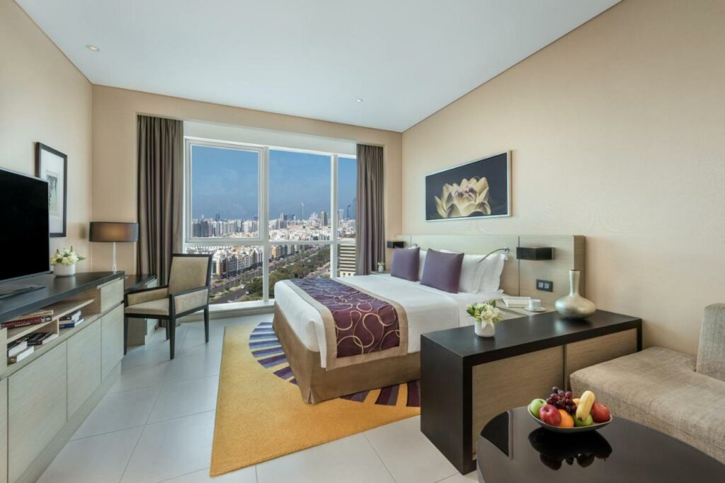 Quarto do Dusit Thani Abu Dhabi. Uma sofá, uma mesa com fruteira e uma cômoda nolado direito, atrás a cama de casal. De frente para a cama, uma cômoda com uma televisão e uma mesa de trabalho. No fundo do quarto, uma parede de vidro com cortinas. 