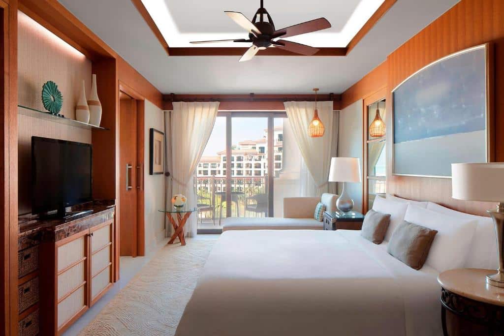 Quarto do The St. Regis Saadiyat Island Resort, Abu Dhabi. Uma cama de casal no lado direito com uma cômoda de cada lado e um abajur de cada lado. De frente para a cama uma raque e uma televisão. No fundo um sofá e uma mesa redonda pequena,e atrás a varanda com duas cadeiras e uma mesa. 