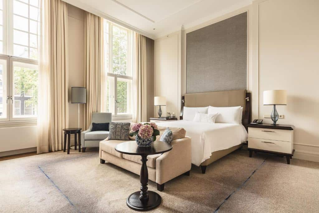 Quarto do Waldorf Astoria Amsterdam espaçoso com uma cama grande, sofá, poltrona, luminárias e janelas altas com cortinas bege.