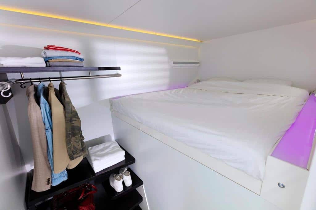 Interior de uma das capsulas do CityHub Amsterdam, de 6 m², com um pequeno cabideiro aoe prateleiras ao lado com roupas dobradas e sapatos, e colchão com travesseiros e ar-condicionado