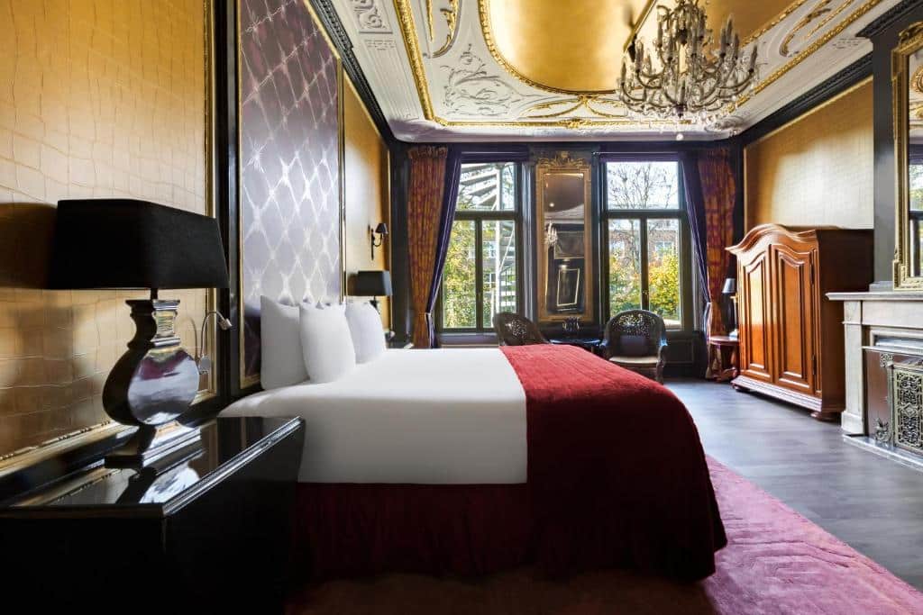 Quarto elegante do The Pavilions Amsterdam, The Toren com teto ornamentado, lustre, cama grande com revestimento vermelho e móveis de madeira vintage ao lado de grandes janelas. Ilustra o post de hotéis em Amsterdam.