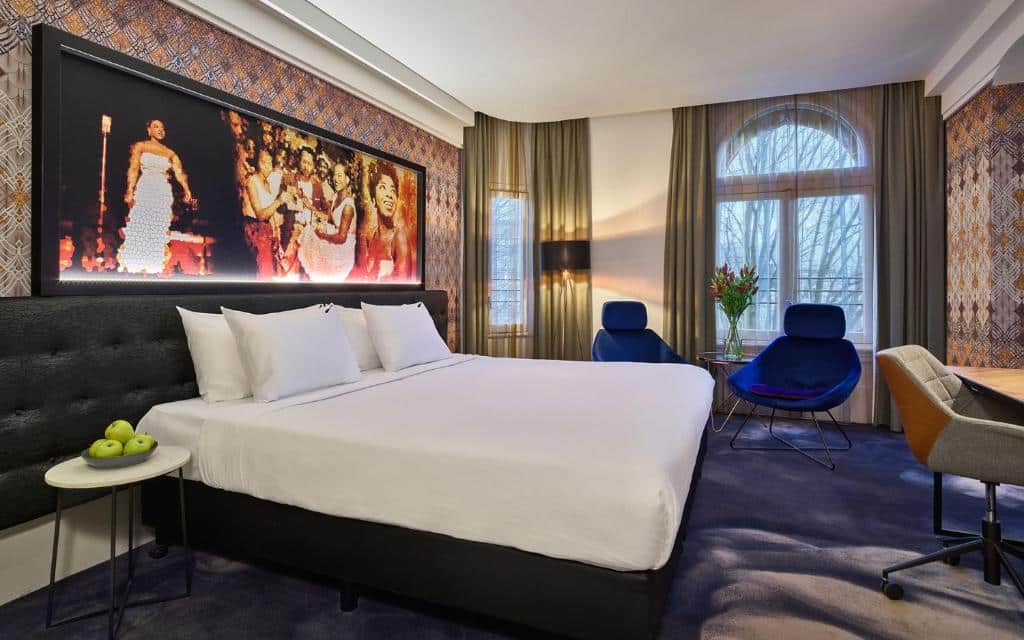 Quarto do Hard Rock Hotel Amsterdam American moderno com uma cama grande, arte na parede acima da cabeceira, área de estar, escrivaninha e uma janela com cortinas transparentes.
