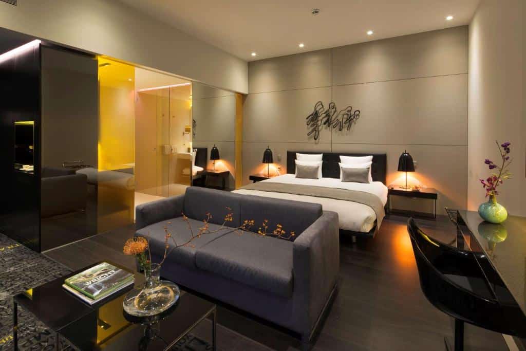 Quarto de hotel art'otel amsterdam moderno com cama king-size, sofá cinza, mesa de vidro e iluminação decorativa; banheiro visível ao fundo.