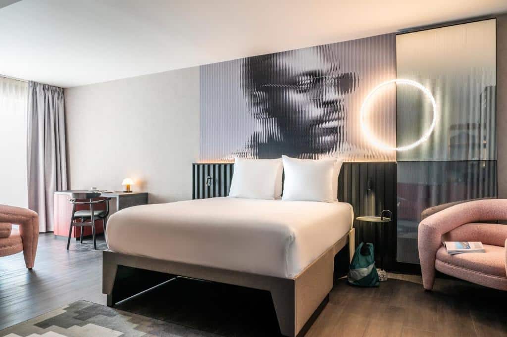 Quarto do Tribe Amsterdam, com cama de casal, uma poltrona e uma mesinha com cadeira. Em cima da cama, há um quadro com o rosto de de um rapaz negro de óculos escuro