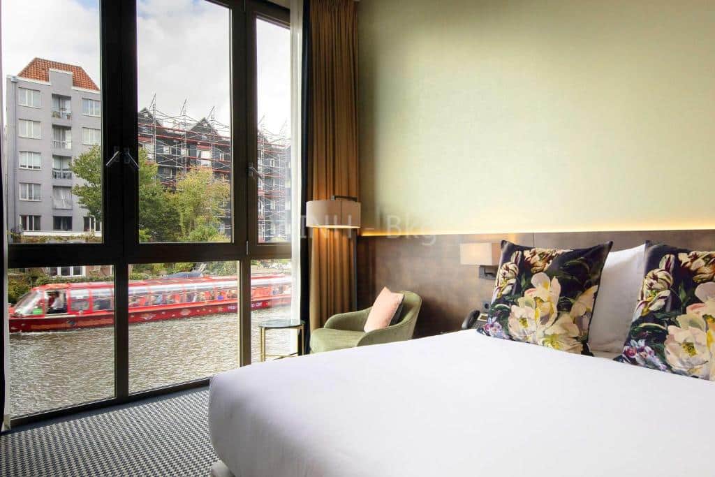 Um quarto do Monet Garden Hotel Amsterdam com uma cama grande, travesseiros florais, uma cadeira verde e uma janela com vista para um canal com um barco vermelho. Ilustra o post de hotéis em Amsterdam.