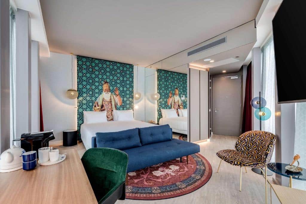 Quarto do nhow Amsterdam Rai, com cama de casal, um sofá na frente azul em cima de um tapete estampado, uma cadeira, e no lado esquerdo tem uma mesa com utensílios para fazer café e uma cadeira verde. Imagem para ilustrar o post de melhores hotéis em Amsterdam