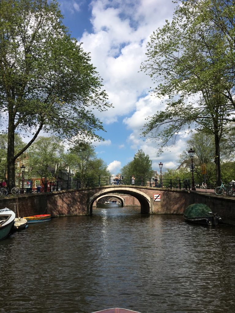 Vista do canal de Amsterdam com margens arborizadas, pequenos barcos e duas pontes de tijolos em arco sob um céu azul com nuvens fofas.