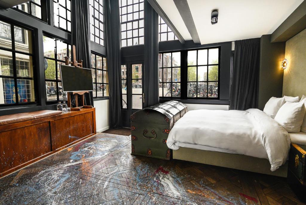 Um quarto moderno do Hotel The Craftsmen com grandes janelas com moldura preta, uma cama branca, um baú de madeira e uma TV em um armário. Ilustra o post de hotéis em Amsterdam.