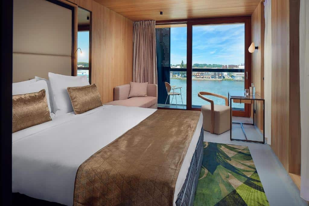 Quarto de hotel moderno do Hotel Jakarta Amsterdam com uma cama grande, área de estar, escrivaninha e varanda com vista para o rio e a paisagem urbana.