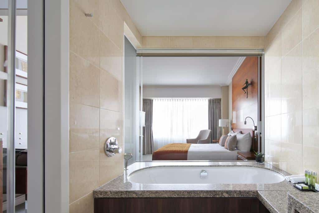 Vista de um banheiro com banheira no para um quarto do Hotel Okura Amsterdam moderno com uma cama, cadeiras e uma grande janela com cortinas.