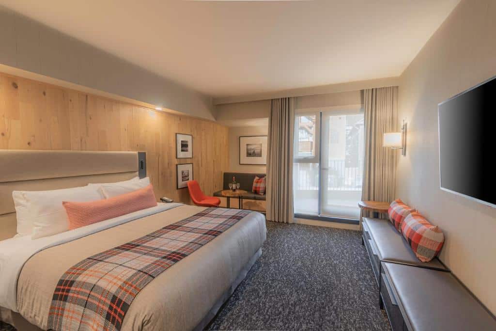 Quarto do Peaks Hotel and Suites com uma cama grande de casal, na frente há alguns lugares para sentar e ao mesmo tempo para guardar objetos com almofadas e em cima na parede uma tv. Ao fundo do lado esquerdo tem uma mesinha redonda com uma garrafa e duas taças, uma cadeira e um banco acoplado na parede com uma almofada e alguns quadros na parede. O chão é de carpete e ao fundo tem uma porta de vidro com uma cortina.