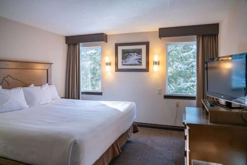 Quarto do The Rundlestone Lodge com uma cama de casal, duas janelas de vidro com cortinas na parede e no meio dessas janelas tem um quadro. Há também uma cômoda de madeira com uma tv em frente a cama.