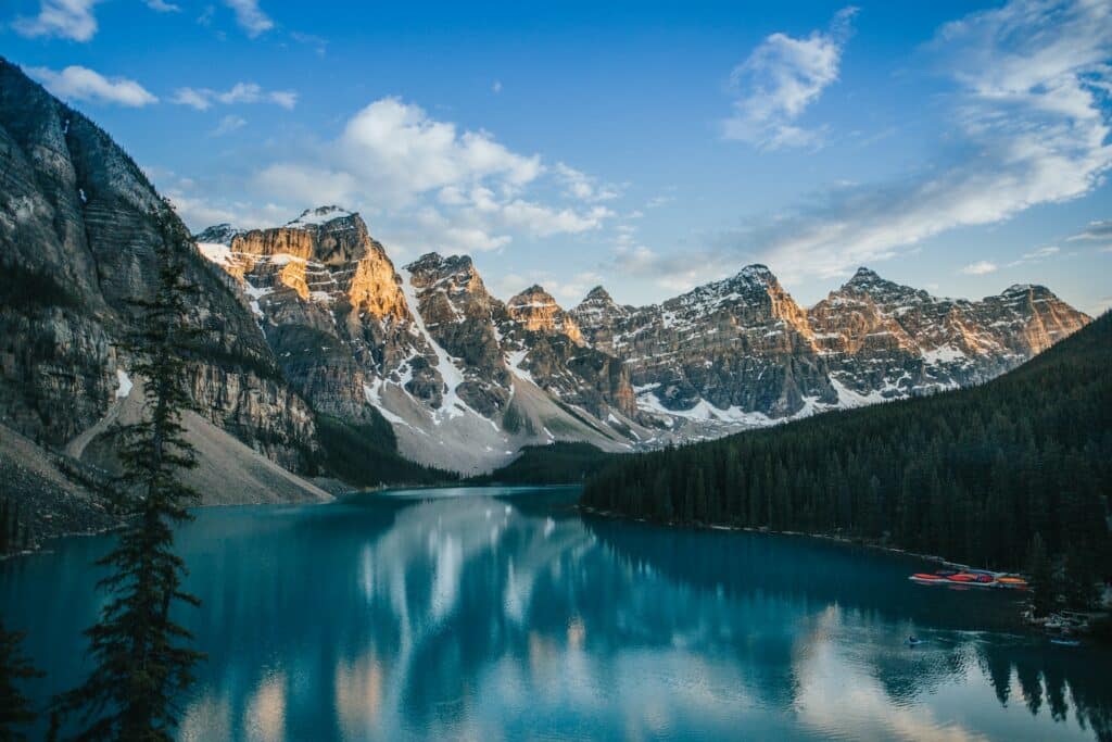 Lago Moraine em Banff com águas azuis, ao lado direito várias árvores verdes e ao lado esquerdo grandes montanhas de pedras cobertas por neve durante o dia, para representar hotéis em Banff.
