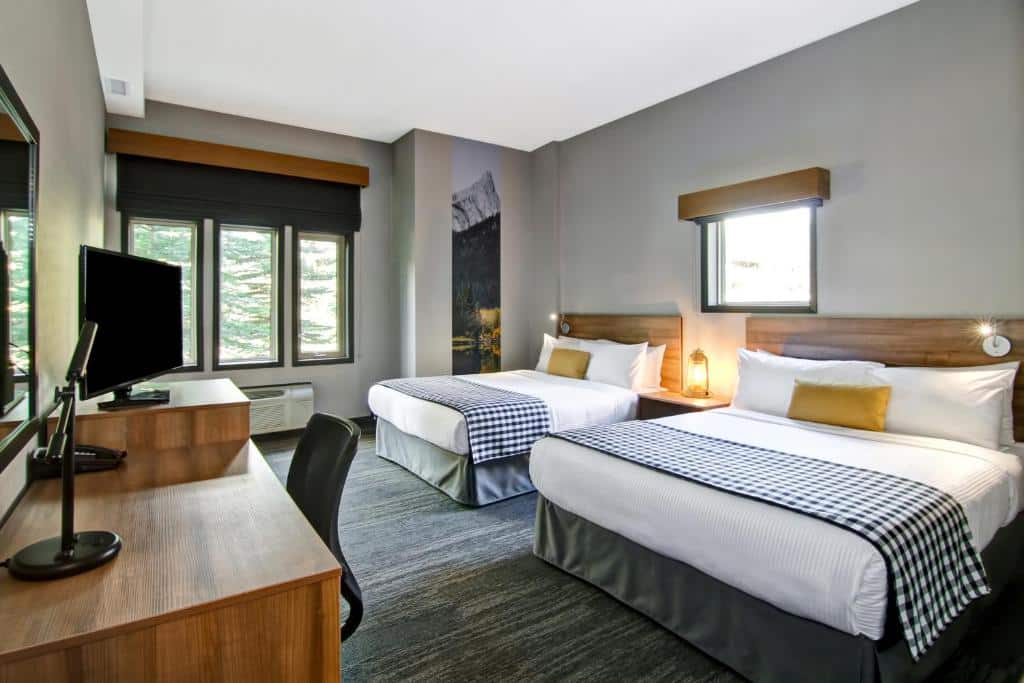 Quarto do Canalta Lodge com duas camas de casal com travesseiros e coberta em cima e uma cabeceira de madeira, no meio das camas há uma mesinha com uma luminária e em cima uma janela quadrada. Em frente as camas tem uma mesa de madeira com uma tv, um telefone, outro aparelho e uma cadeira, já ao fundo tem três janelas retangulares.