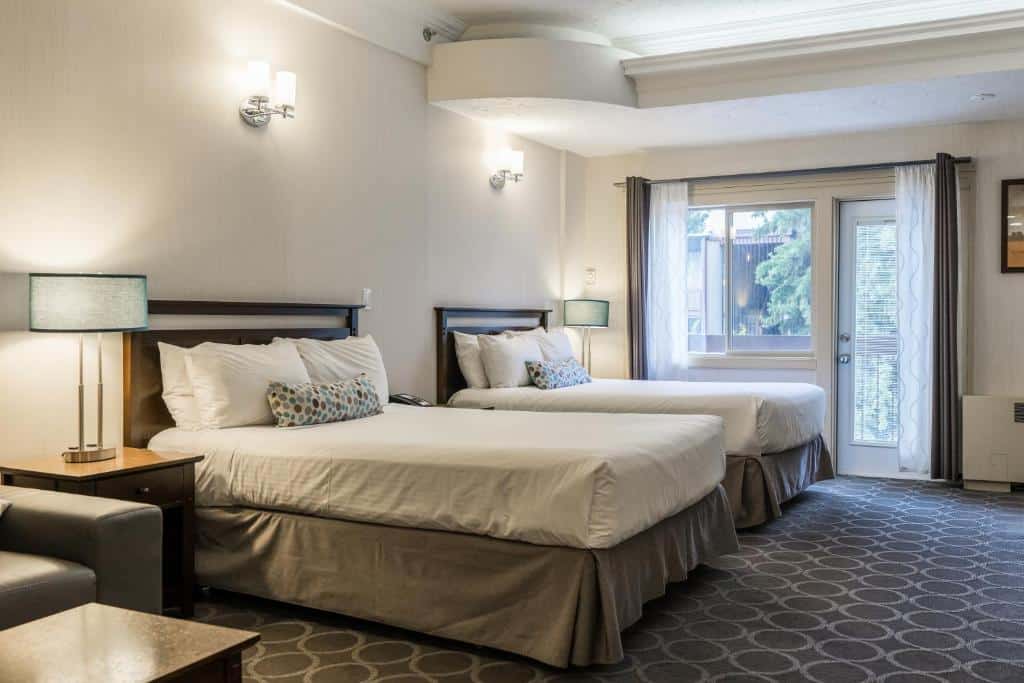 Quarto do High Country Inn com duas camas de casal, em cada lado da cama há uma mesinha de madeira com um abajur e ao lado esquerdo da foto tem uma poltrona com um banquinho. O chão é de carpete e ao fundo tem uma porta e uma janela de vidro com uma cortina.