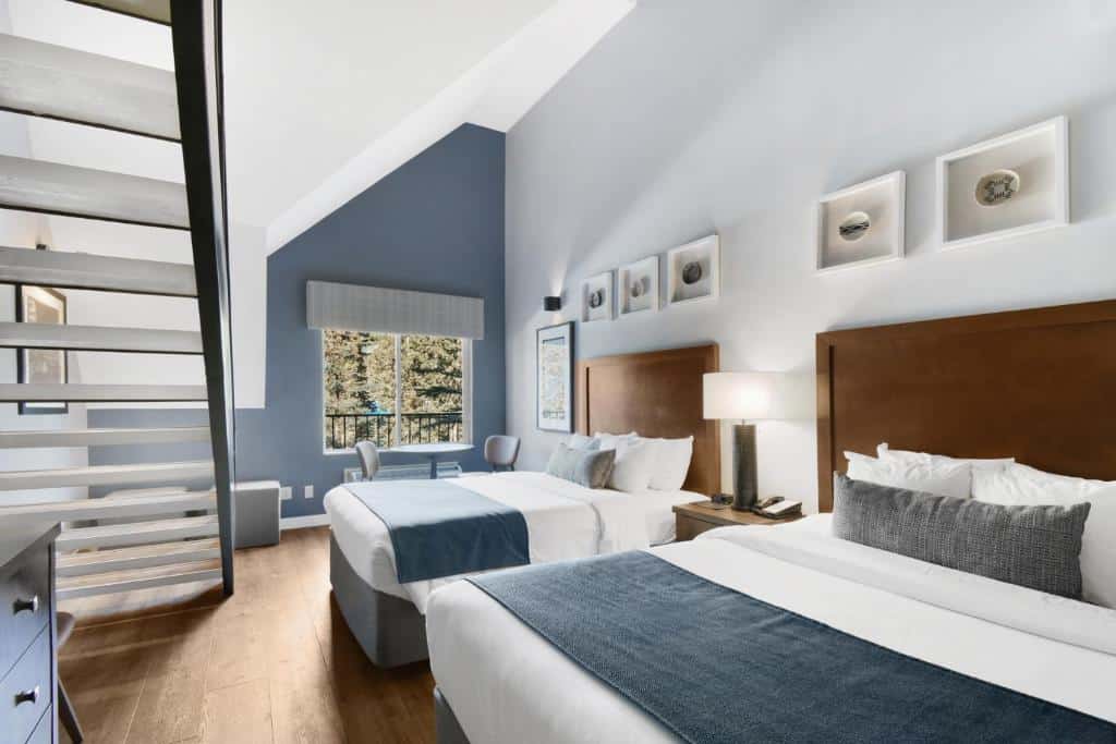 Quarto do hotel Banff Inn com duas camas de casal com cabeceiras de madeira, no meio das camas há uma mesinha com um abajur e vários quadros na parede. No fundo do quarto tem duas cadeiras, uma janela de vidro e mais para o canto esquerdo tem uma escada.