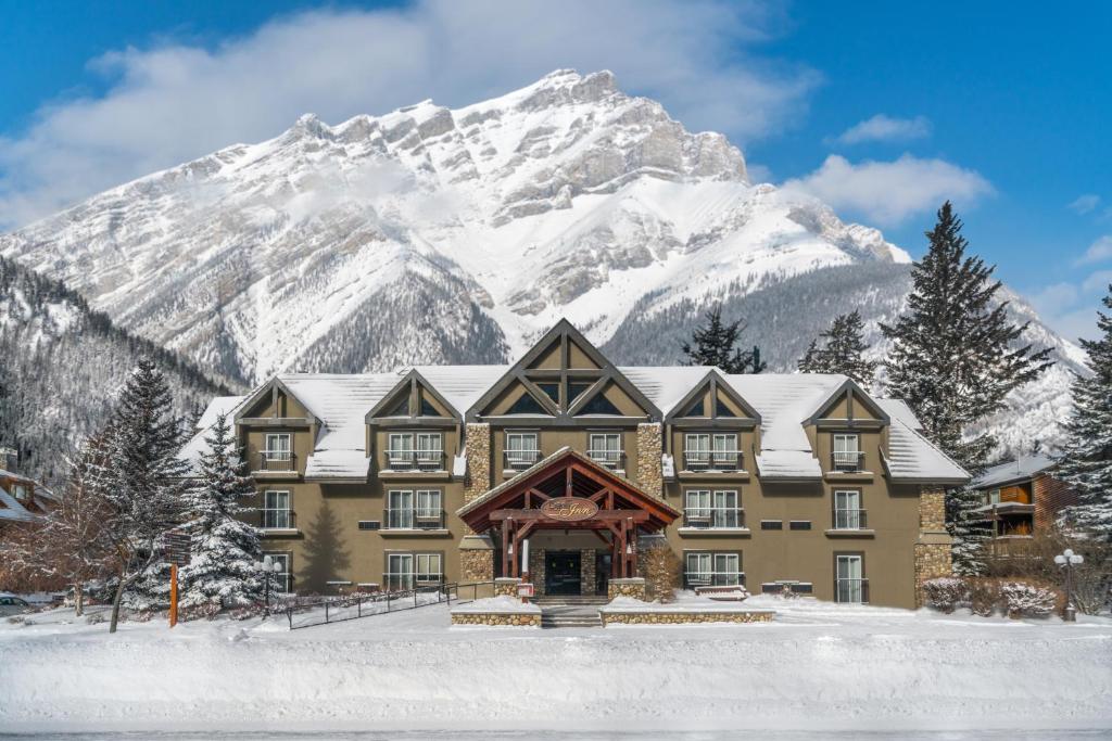 Fachada do hotel Banff Inn na cor marrom com três andares e telhados pontudos. Em volta tem algumas árvores cobertas por neve e ao fundo uma montanha coberta por neve também, para representar hotéis em Banff.