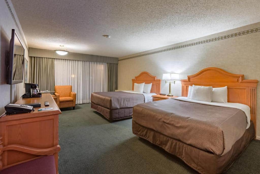 Quarto do Banff Park Lodge com duas camas de casal separadas por uma mesinha de madeira com um abajur. Na frente das camas há uam mesa maior de madeira com alguns objetos e em cima na parede uma tv. Ao fundo tem uma poltrona amarela e atrás dela uma cortina branca e cinza.