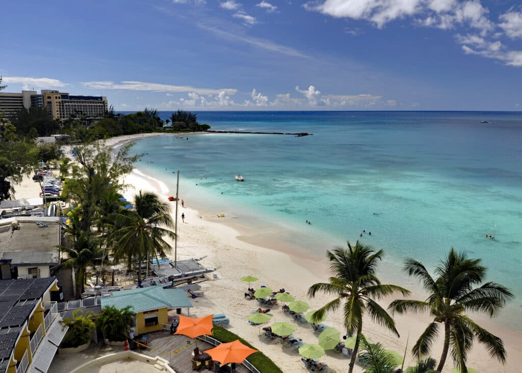 Vista de cima de Carlisle Bay, em Barbados, com uma água cristalina palmeiras e uma orla com guarda-sóis.