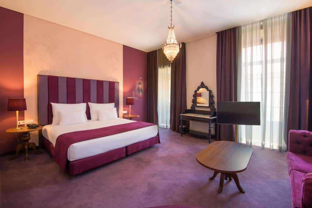 Um quarto no Vila Gale Collection Braga com uma cama king-size, decoração roxa, lustre, escrivaninha com espelho, TV, duas mesas laterais e janelas grandes com cortinas, para representar hotéis em Braga.