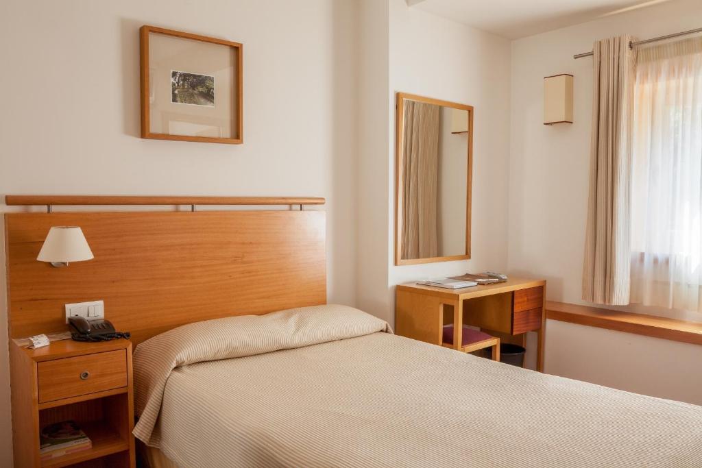 Um pequeno quarto no Hotel do Lago com uma cama de solteiro, cabeceira de madeira, escrivaninha, espelho, janela com cortinas e arte emoldurada, para representar hotéis em Braga.