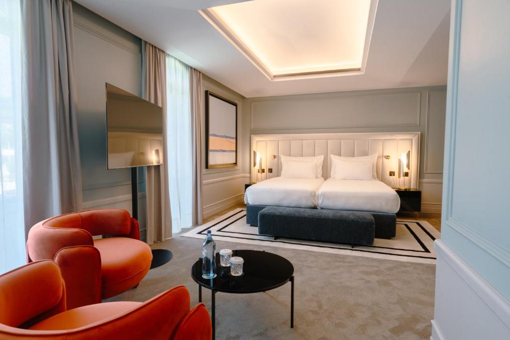 Quarto de hotel moderno com duas camas, uma TV na parede, duas cadeiras laranjas, uma mesa redonda e janelas grandes com cortinas.