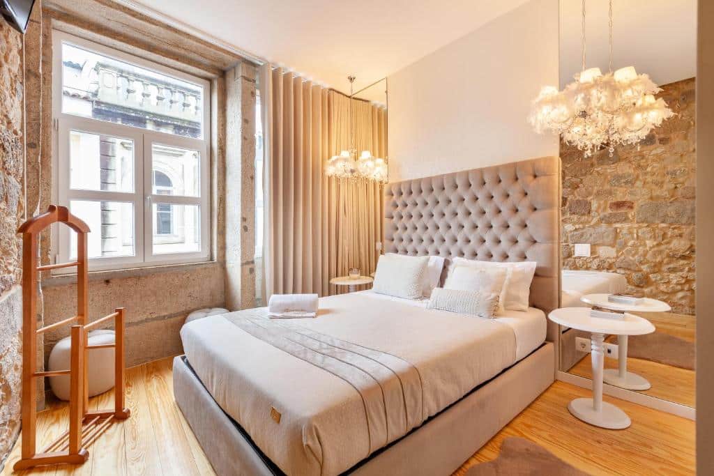 Uma cama bem arrumada em um quarto bem iluminado com paredes de pedra, uma janela, um espelho e luminárias modernas, no hotel Berço das Mordomias, para representar hotéis em Braga.