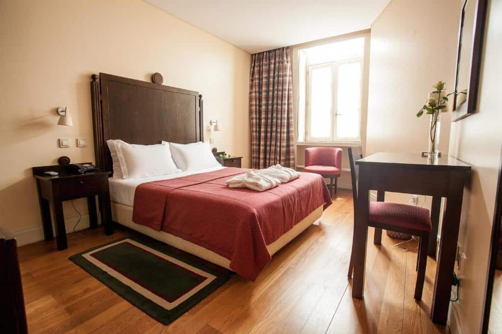 Um quarto no Hotel Bracara Augusta arrumado com uma cama de casal, colcha vermelha, piso de madeira, escrivaninha, cadeira, janela e mesas de cabeceira, para representar hotéis em Braga.