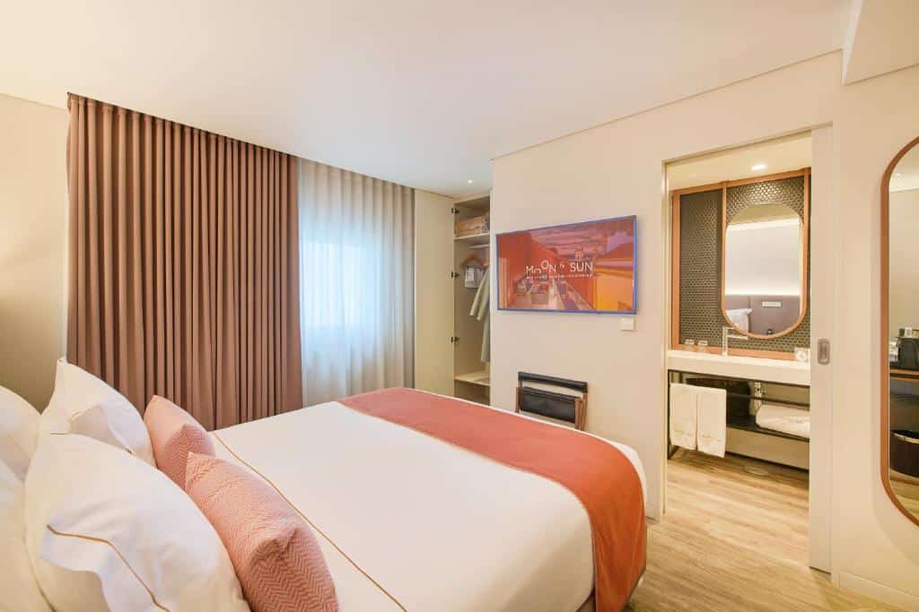 Um quarto moderno no Hotel Moon & Sun Braga com uma cama bem arrumada, TV na parede, janela com cortinas e uma área de banheiro visível, para representar hotéis em Braga.