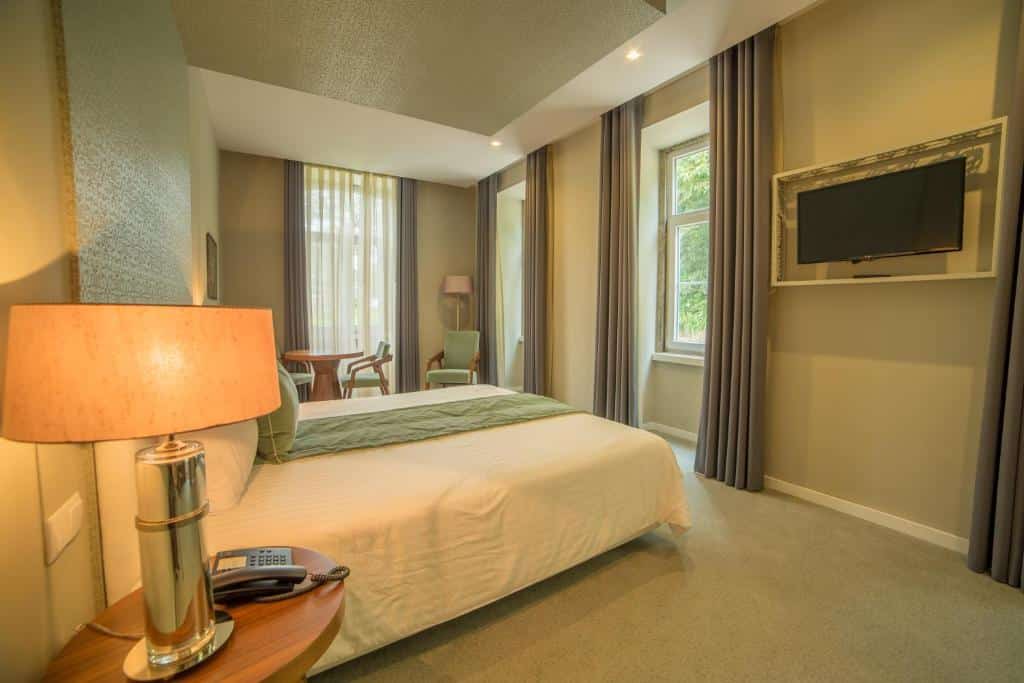 Um quarto no Hotel do Parque com uma cama de casal, mesa de cabeceira, abajur, TV na parede, cadeiras e janelas grandes com cortinas, para representar hotéis em Braga.
