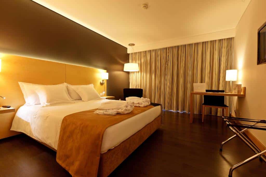 Quarto moderno no Hotel Mercure Braga Centro com uma cama de casal, mesas de cabeceira, escrivaninha com laptop, cadeira e janela grande com cortinas fechadas, para representar hotéis em Braga.