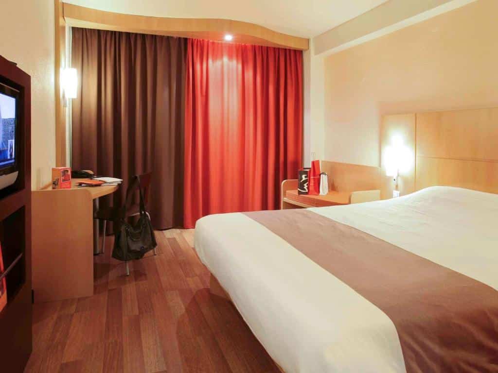 Um quarto no Hotel Ibis Braga com uma cama de casal, piso de madeira, uma pequena escrivaninha, cadeira, TV e cortinas vermelhas e marrons fechadas, para representar hotéis em Braga.
