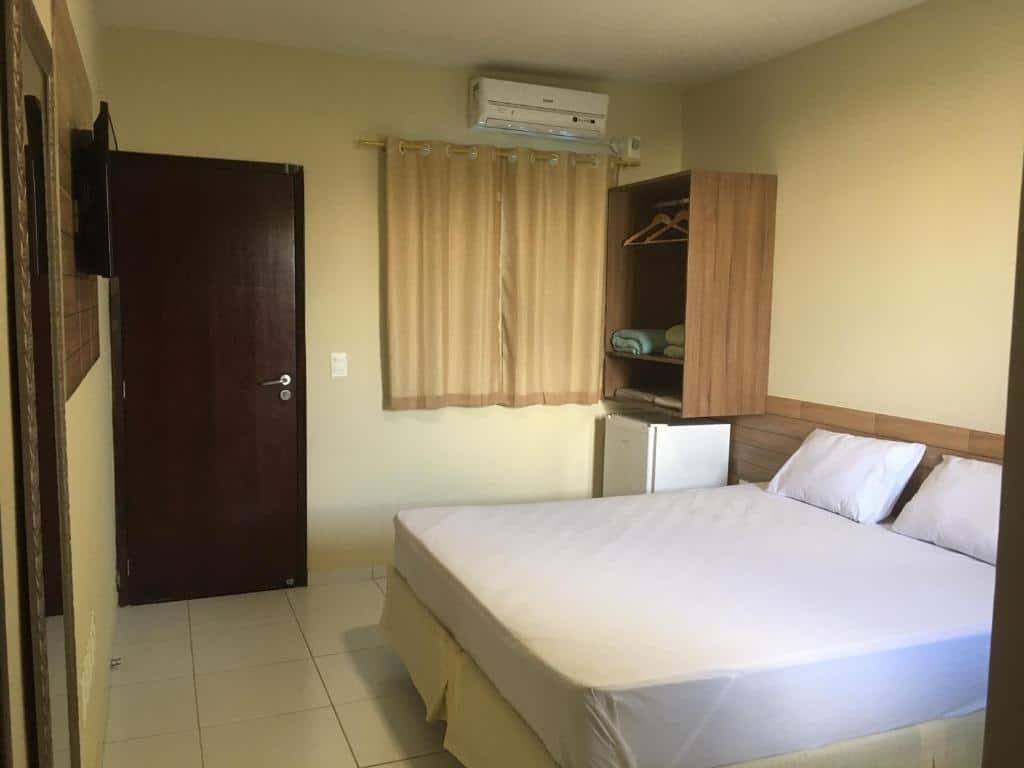 Quarto da Pousada Sumaré. A cama está no lado esquerdo e em frente a cama há um espelho e uma TV presa a parede. Ao fundo há uma porta, cortina, frigobar e pequeno armário em cima do frigobar. Imagem para ilustrar o post hotéis em Caruaru.