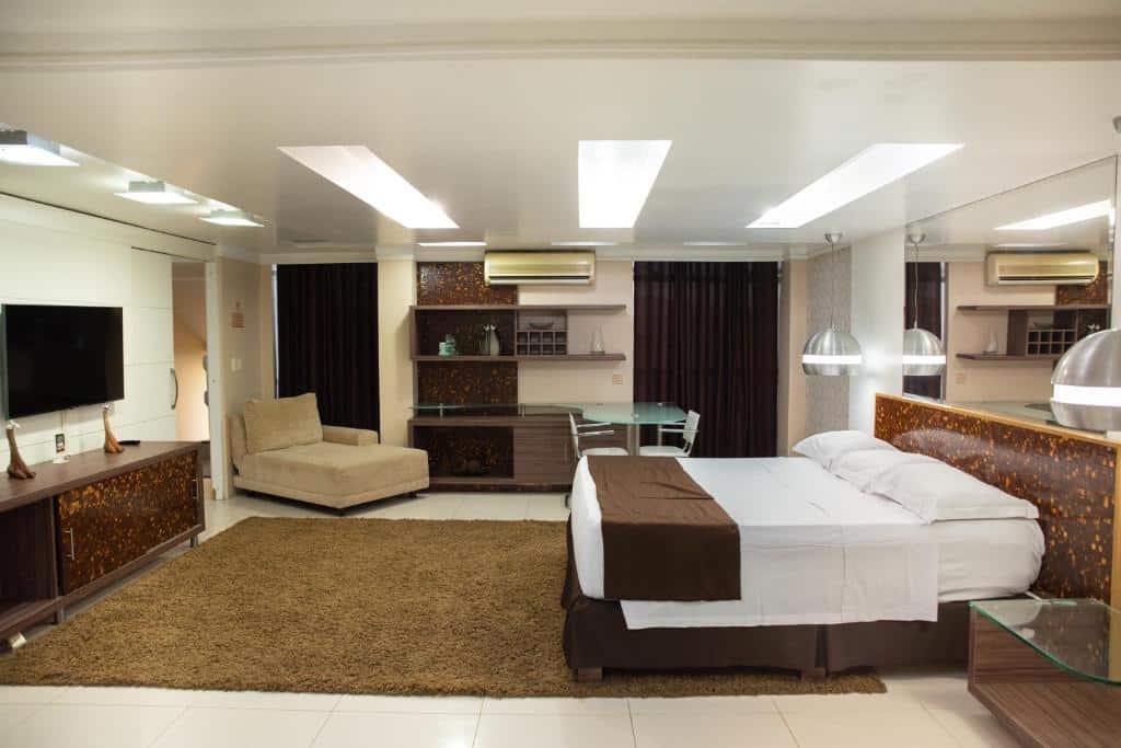 Quarto do Hotel Village Premium Caruaru. O quarto é bem amplo, a cama está encostada na parede direita com um espelho em cima. Já na parte esquerda há uma cômoda com uma TV em cima presa a parede. Ao fundo está uma grande mesa com duas cadeiras, uma poltrona, prateleiras presas a parede e duas grandes cortinas. Imagem para ilustrar o post hotéis em Caruaru.