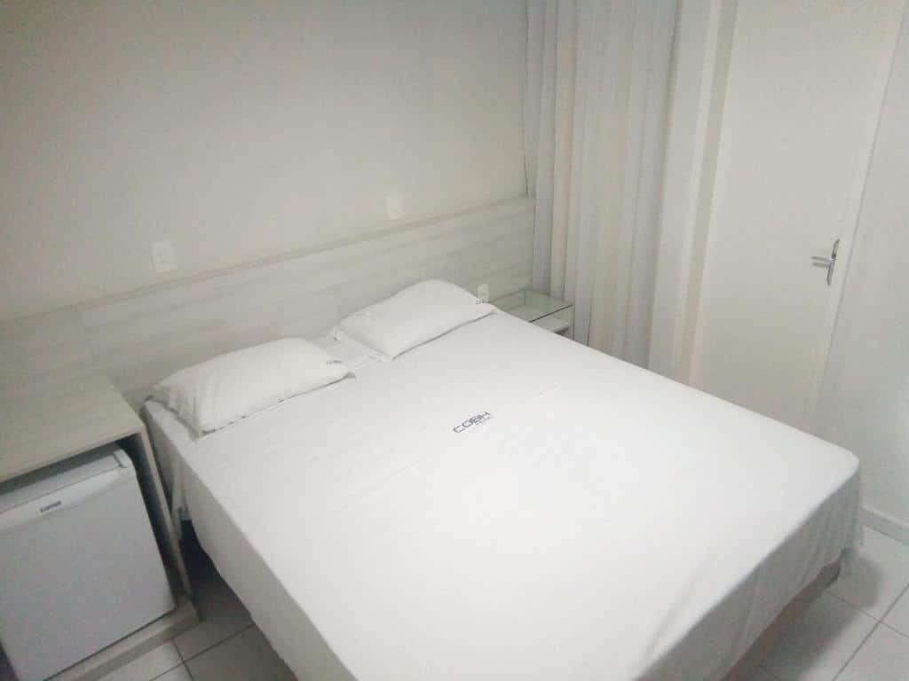Quarto do COBH Hotel. A cama está centralizada, no lado esquerdo da cama há um frigobar, no lado direito há uma pequena prateleira. Também na parede direita há uma porta e uma cortina. Imagem para ilustrar o post hotéis em Caruaru.