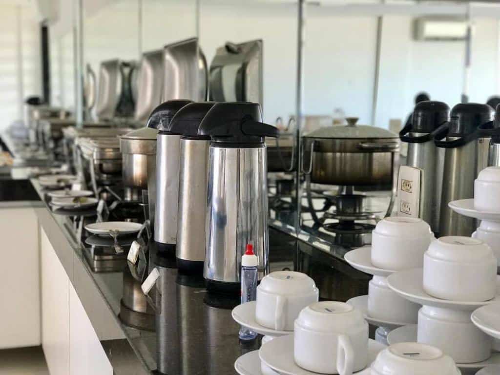 Área para café da manhã do COBH Hotel. Em cima de uma bancada de granito estão garrafas térmicas, xícaras e panelas.