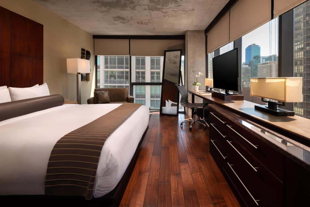 Quarto do Eurostars Magnificent Mile com cama de casal ampla, cômoda em frente a cama com TV e mesa de trabalho com cadeira ao lado, do lado esquerdo, sofá cinza ao lado de um espelho grande.
