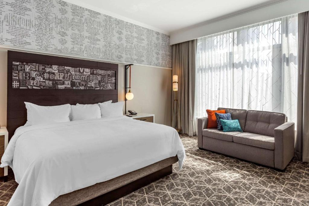 Quarto do Hotel Blake, an Ascend Hotel Collection Member, com cama de casal, uma cômoda de madeira do lado esquerdo perto de uma sofá de dois lugares cinza em frente a janelas com cortinas brancas e cinzas.
