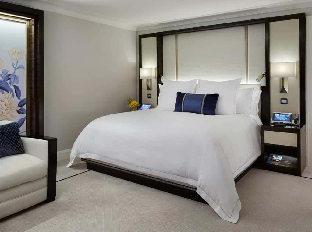 Quarto moderno no The Peninsula Chicago com uma cama grande, roupa de cama branca, duas mesas de cabeceira, luminárias de parede e uma poltrona branca.