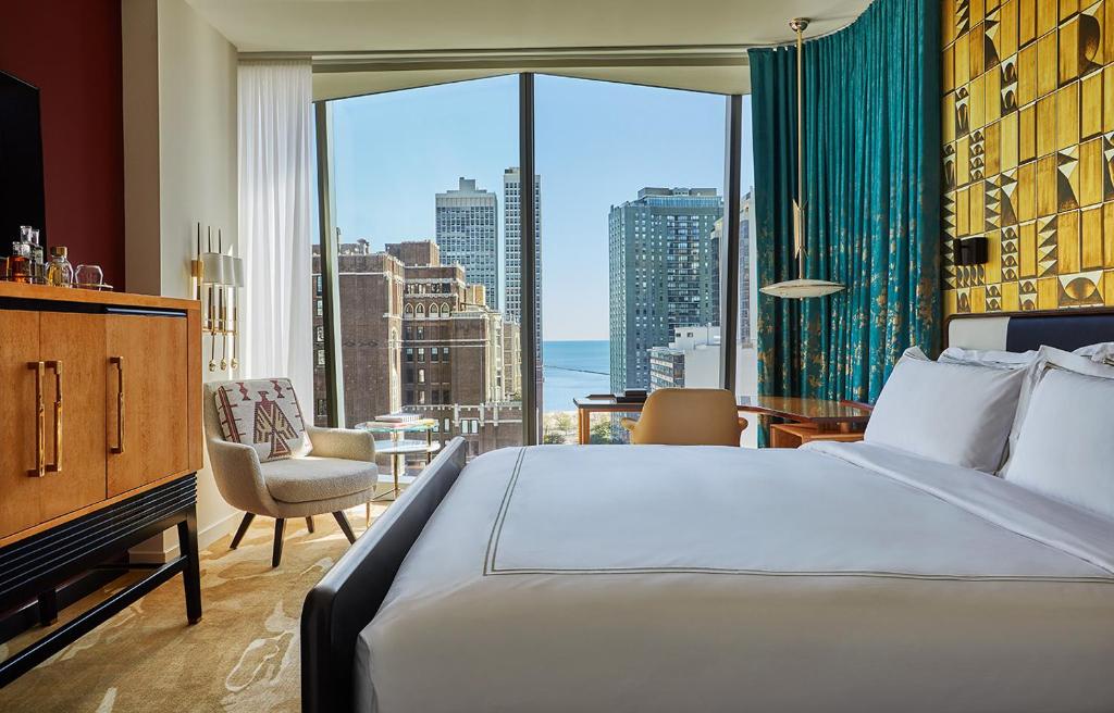 Quarto do Viceroy Chicago com cama do lado direito, em frente a cama uma cômoda de madeira com TV, uma poltrona branca e do lado esquerdo mesa de trabalho em frente a janelas panorâmicas com vista para a cidade, para representar hotéis em Chicago.