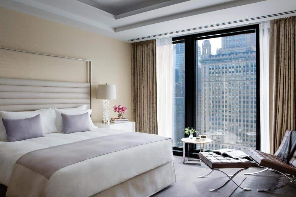 Quarto do The Langham com cama de casal, uma cômoda branca com luminária do lado esquerdo, com poltrona de couro e mesa redonda perto das janelas do lado direito.