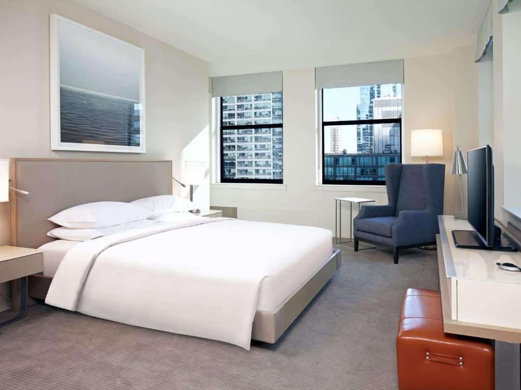 Quarto do Hyatt Centric The Loop com cama de casal, duas cômodas com luminárias em frente a cama uma cômoda com TV e do lado esquerdo do quarto poltrona azul do lado das janelas.