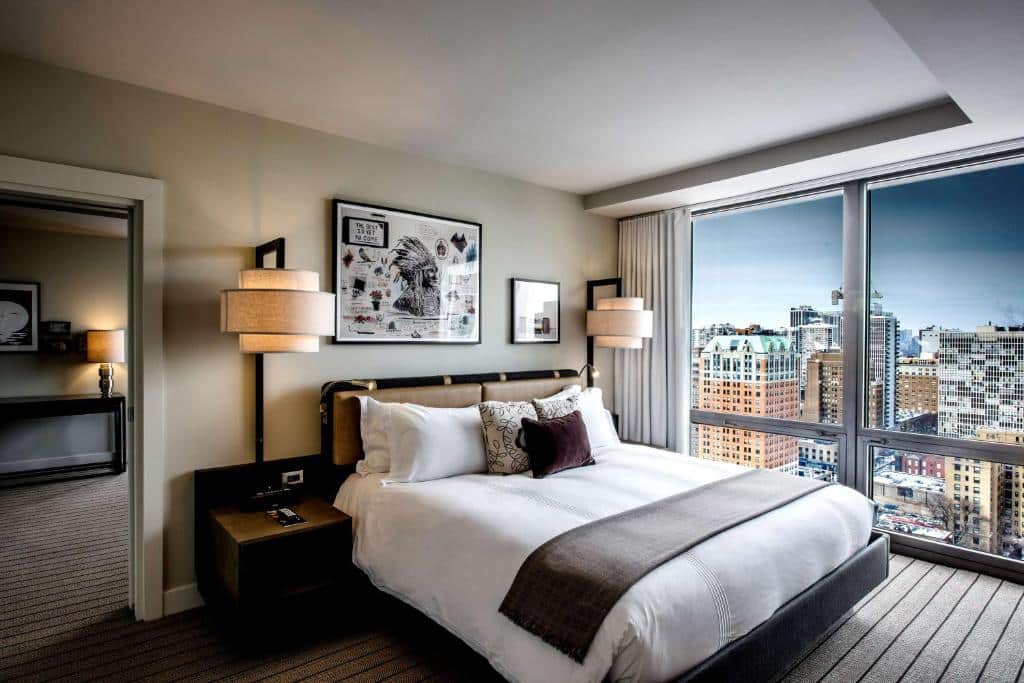 Quarto do Thompson Chicago com cama de casal, duas cômodas ao lado da cama, com luminárias e janelas panorâmicas com cortinas brancas.