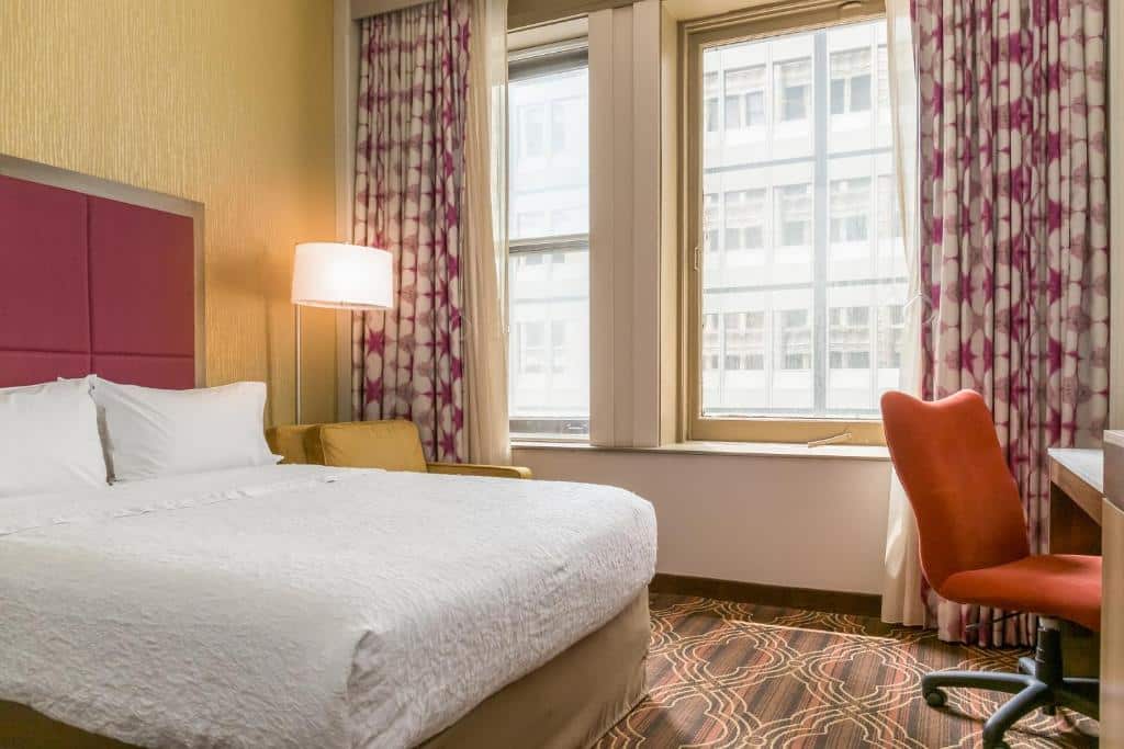 Quarto do Hampton Inn Majestic Chicago Theatre District com cama de casal, poltrona do lado esquerdo com luminária e em frente mesa de trabalho com cadeira laranja.