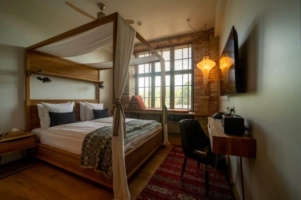 Quarto do hotel Bryggen Guldsmeden. Há uma cama de casal com um pergolado, uma luminária com luz amarela, mesa com cadeira, televisão e uma janela com um conchonete para se deitar. Foto para ilustrar post sobre hotéis em Copenhague.