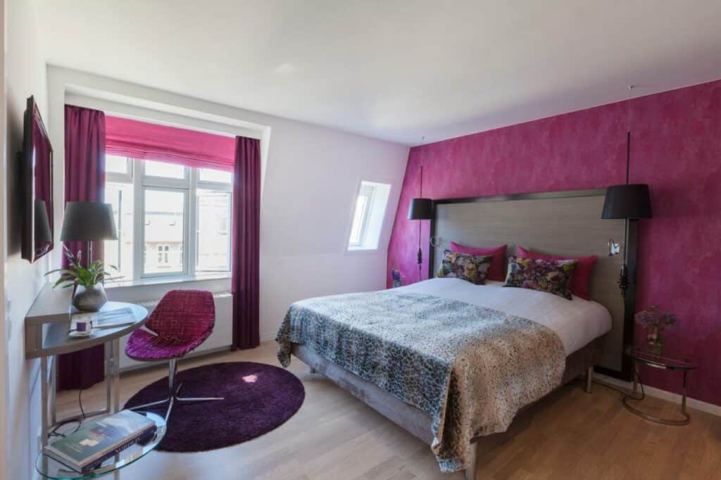 Quarto do hotel Andersen Boutique Hotel com uma cama de casal, televisão e uma mesinha com cadeira. Uma parede do quarto e as cortinas são rosas. 