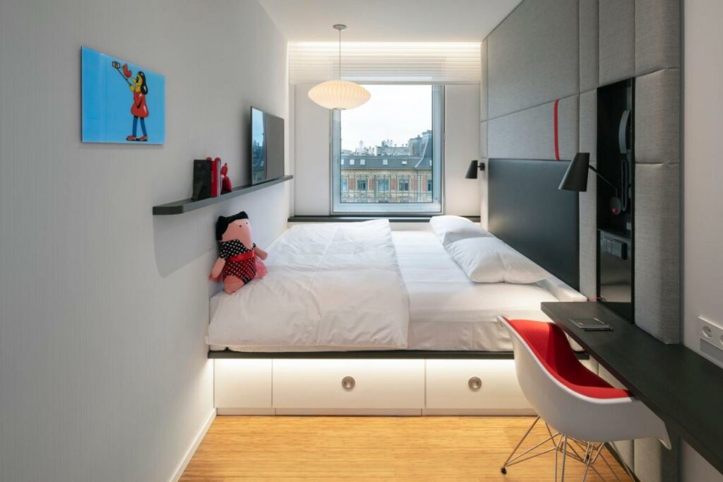 Quarto do citizenM Copenhagen Radhuspladsen com uma cama grande de casal com cabeceira toda acolchoada, uma prateleira sustentando uma televisão e uma mesa estreita com cadeira. Há também uma janela. Foto para ilustrar post sobre hotéis em Copenhague.