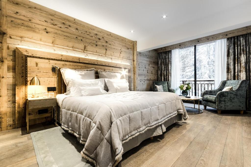 Quarto do Hôtel La Sivolière. Há uma cama de casal encostada na parede e bem no centro do ambiente. Do lado esquerdo existe uma mesinha com abajur e, do lado direito, duas poltronas e uma mesa em meio a elas. Atrás delas há uma janela com vista para a neve. Foto para ilustrar post sobte hotéis em Courchevel.