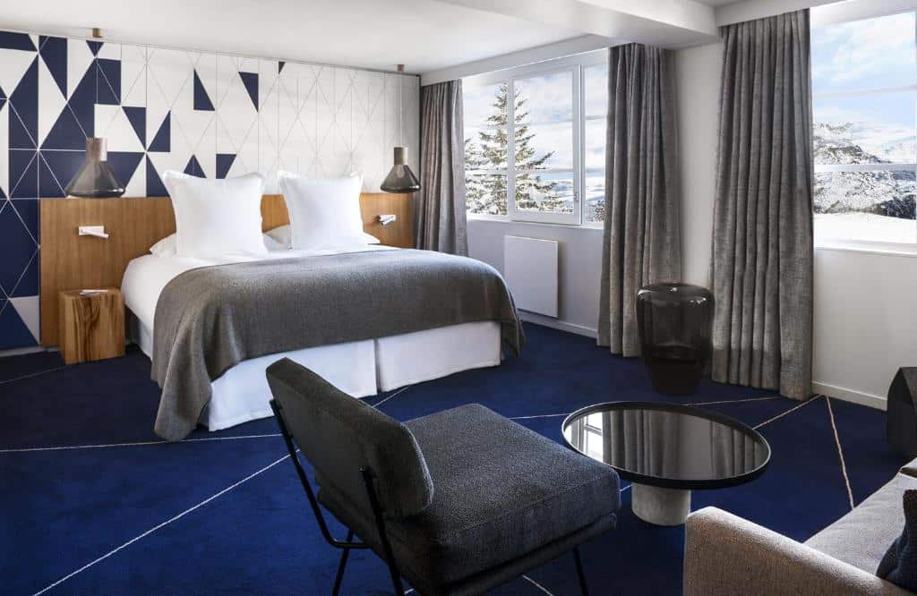 Quarto do hotel White 1921 Courchevel. Ele tem 34 m² e a cama está encostada na parede e no centro do ambiente. Ao lado dela existem janelas que dão vista para as montanhas e para as árvores e, em frente, há um sofá, uma cadeira e uma mesinha de centro redonda. Foto para ilustrar post sobre hotéis em Courchevel.