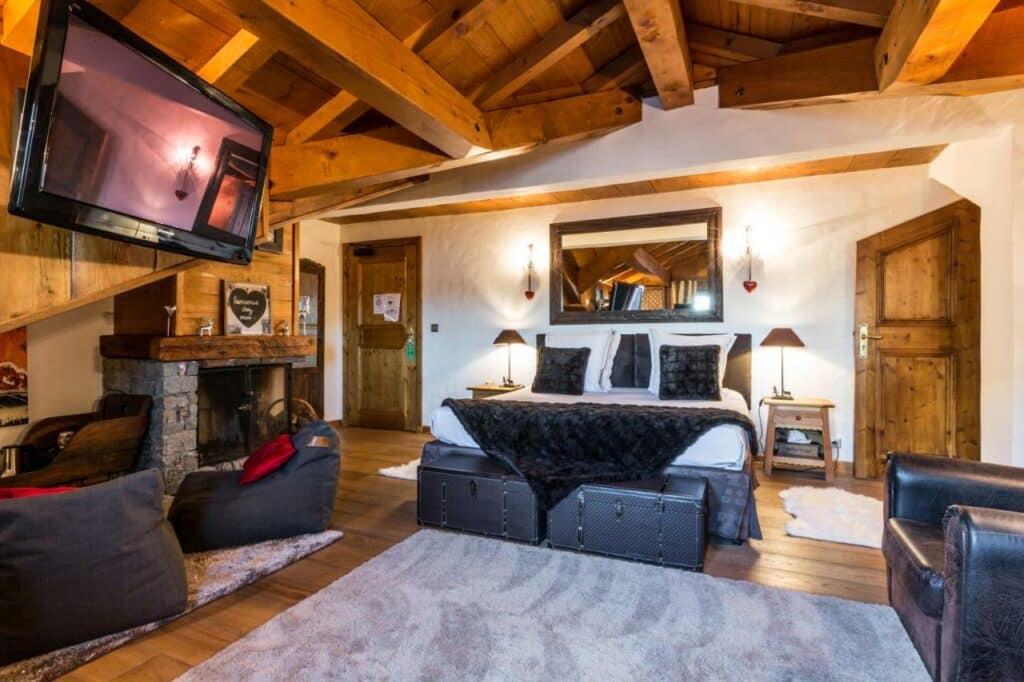 Quarto do hotel Les Monts Charvin com um cama de casal encostada na parede e bem no centro. Ao pé da cama existem dois baús. Do lado esquerdo é possível ver uma lareira, dois pufes e uma televisão. Do lado direito há uma poltrona virada para a tv. Foto para ilustrar post sobre hotéis em Courchevel. 